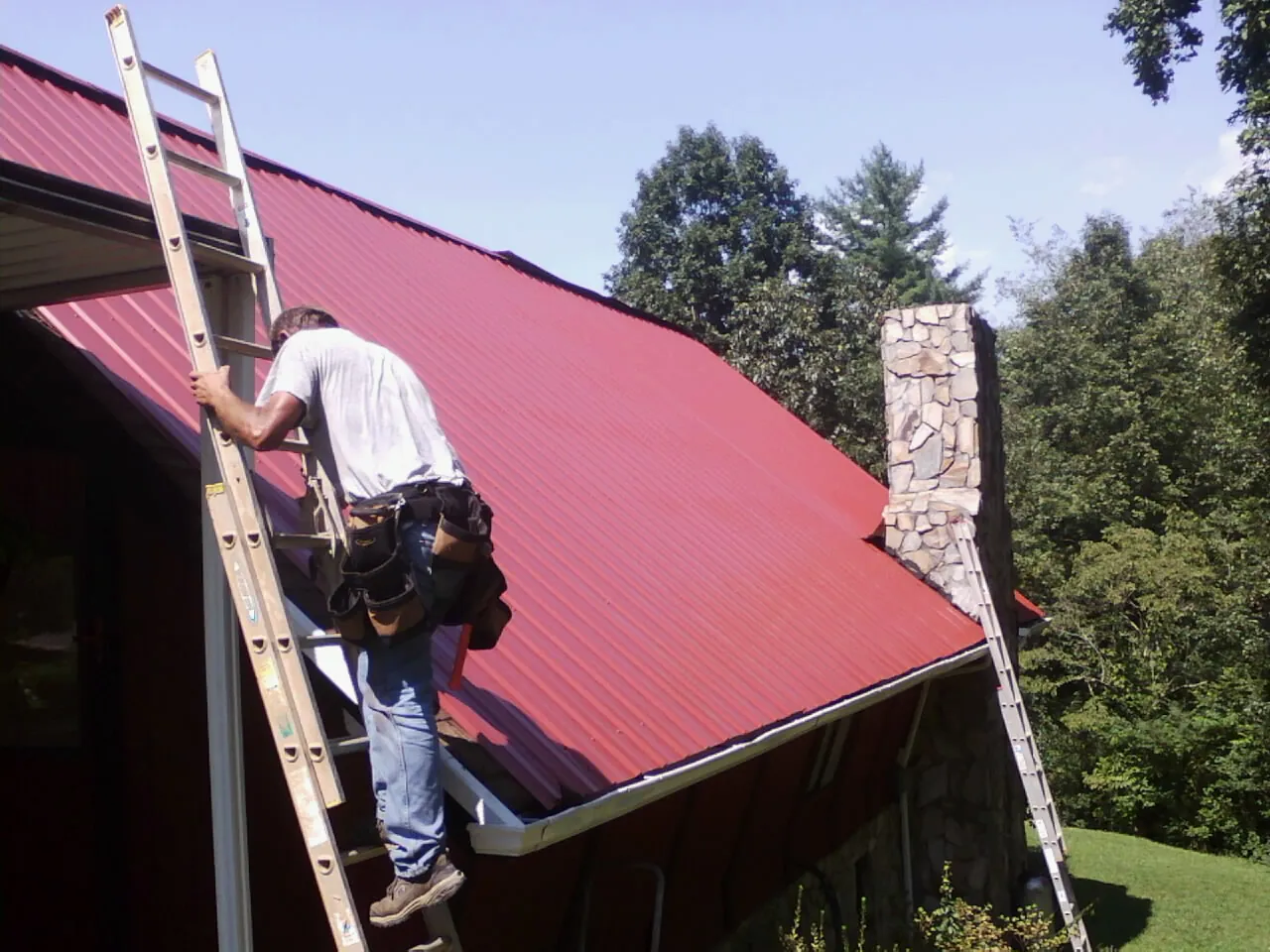 Roofer on a ladder beside a red metal roof and stone chimney — Soffit & Fascia Repair by Roofing Brothers in Idanha